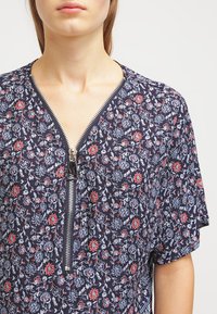 Laivastonsininen kukallinen paita lyhyillä hihoilla, jossa on edessä vetoketju ja monimutkaisia punaisia ja sinisiä kukkakuvioita pehmeällä kankaalla.