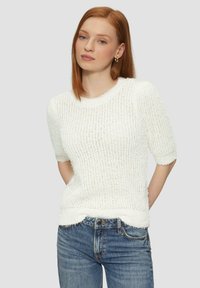 Biały sweter o fakturowanym dzianinowym wykończeniu z krótkimi rękawami, z okrągłym dekoltem i postrzępionym dołem. Noszony z niebieskimi dżinsami.