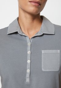 Grå polotröja med krage, fem knappar framtill och bröstficka på vänster sida. Mjuk tygkvalitet med en slät textur.