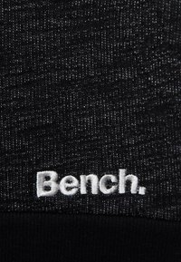 Tissu noir avec un motif texturé ; comporte un logo "Bench." brodé en blanc en bas. Fini doux, matériau extensible.