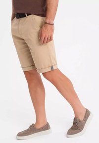 Mand iført beige shorts til knæene, brun bælte, brun skjorte, brun armbånd og taupe bådsko på hvid baggrund.