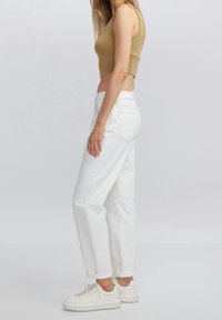 Donna con top corto beige, jeans bianchi a vita alta con risvolti, e sneakers bianche, in piedi di lato contro uno sfondo semplice.