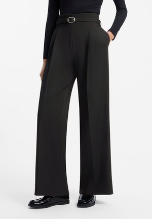 Person trägt schwarze, weit geschnittene Hose mit hohem Bund und Gürtel, schwarzes Langarmshirt und schwarze glänzende Loafers, steht mit einer Hand in der Tasche.