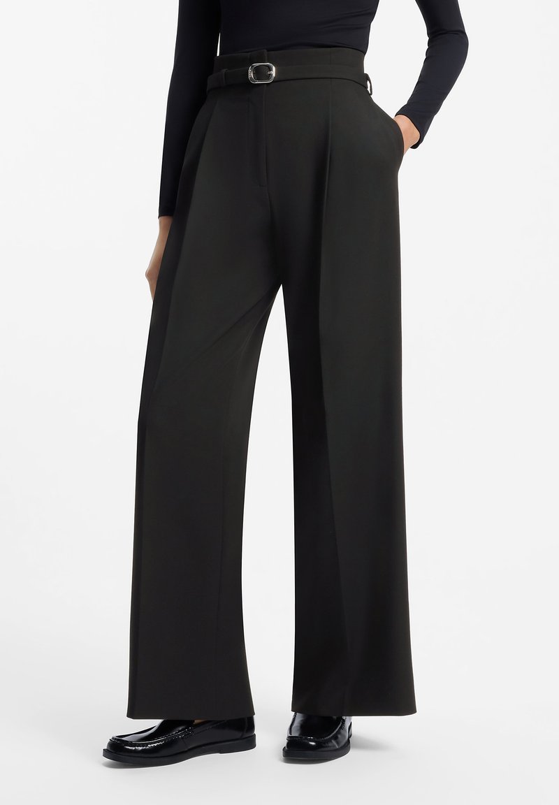 Personne portant un pantalon noir taille haute à jambes larges avec une ceinture, un haut noir à manches longues et des mocassins noirs brillants, debout avec une main dans la poche.