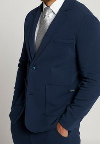 Muž v námořnickém blejzru se dvěma předními náprsními kapsami, bílé košili a světlým šedým vzorovaným kravatě, s rukama v kapsách kalhot.