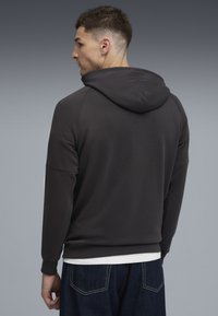 Mand der vender til venstre, iført en mørkegrå hættejakke, hvid undertrøje og mørkeblå jeans på en ensfarvet grå baggrund.