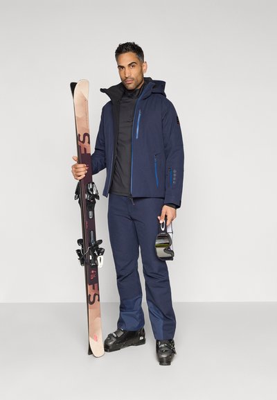 Námořnická lyžařská bunda s modrými detaily a kapsami na zip, doplněná o odpovídající kalhoty. Drží hnědé lyže a černé lyžařské brýle.