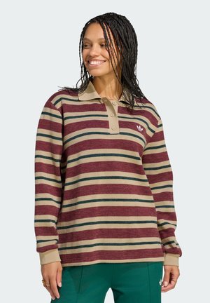 Smiling woman with braided hair wears a long-sleeve striped polo shirt in maroon, beige, and green with green pants against a plain background.