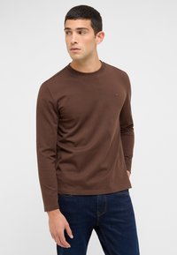 Braunes Langarmshirt mit Rundhalsausschnitt aus Baumwolle, das ein dezentes Logo auf der Brust zeigt, kombiniert mit dunkelblauen Denim-Jeans.