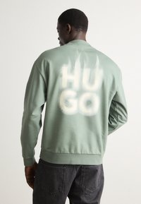 Zöld, hosszú ujjú pulóver, kerek nyakkivágással. A hátulján fehér színnel, texturált hatással van feliratozva a "HUGO". Klasszikus fazon.