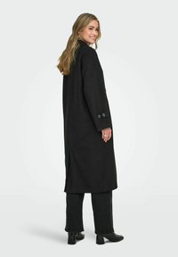 Ilgas juodas vilnos mišinio paltas, su aukštu apykakliu, šoninėmis angomis ir mygtukuose užsegamomis rankovėmis, derinamas su juodais džinsais ir kulkšninių botais.