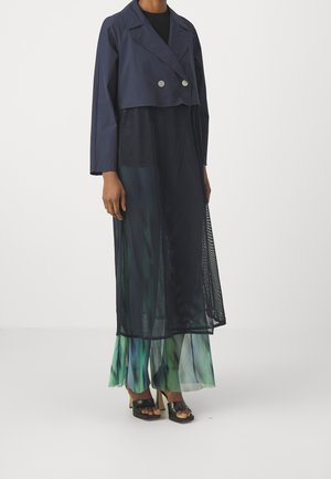 Persoon draagt een marineblauw cropped dubbelreeks jasje over een doorschijnende zwarte lange jurk, met groen-blauwe wijde broekspijpen en zwarte sandalen met hak.