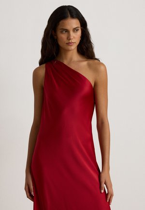 Femme aux longs cheveux foncés portant une robe rouge en satin à une épaule, debout devant un fond gris clair uni.