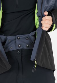 Vattentät jacka med ett mörkt yttre lager och ljusgröna detaljer. Inkluderar en mudd med dragkedja, justerbara manschetter och tryckknappsstängning vid midjan.