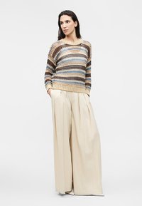 Femme portant un pull en tricot rayé multicolore et un pantalon beige plissé à jambes larges, les mains dans les poches, debout devant un fond uni.