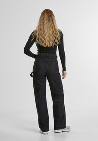 Pantaloni neri a vita alta, ampi, con tasche laterali, realizzati in denim testurizzato e dettagli con cintura. Indossati con un top aderente a maniche lunghe.