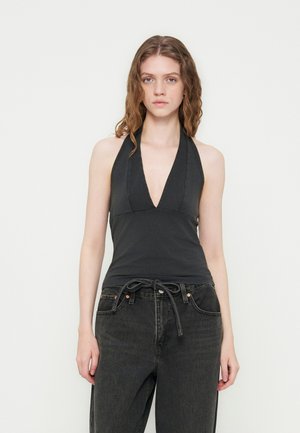 Jeune femme aux cheveux ondulés portant un haut noir sans manches à col halter et un jean large gris foncé avec une ceinture nouée.