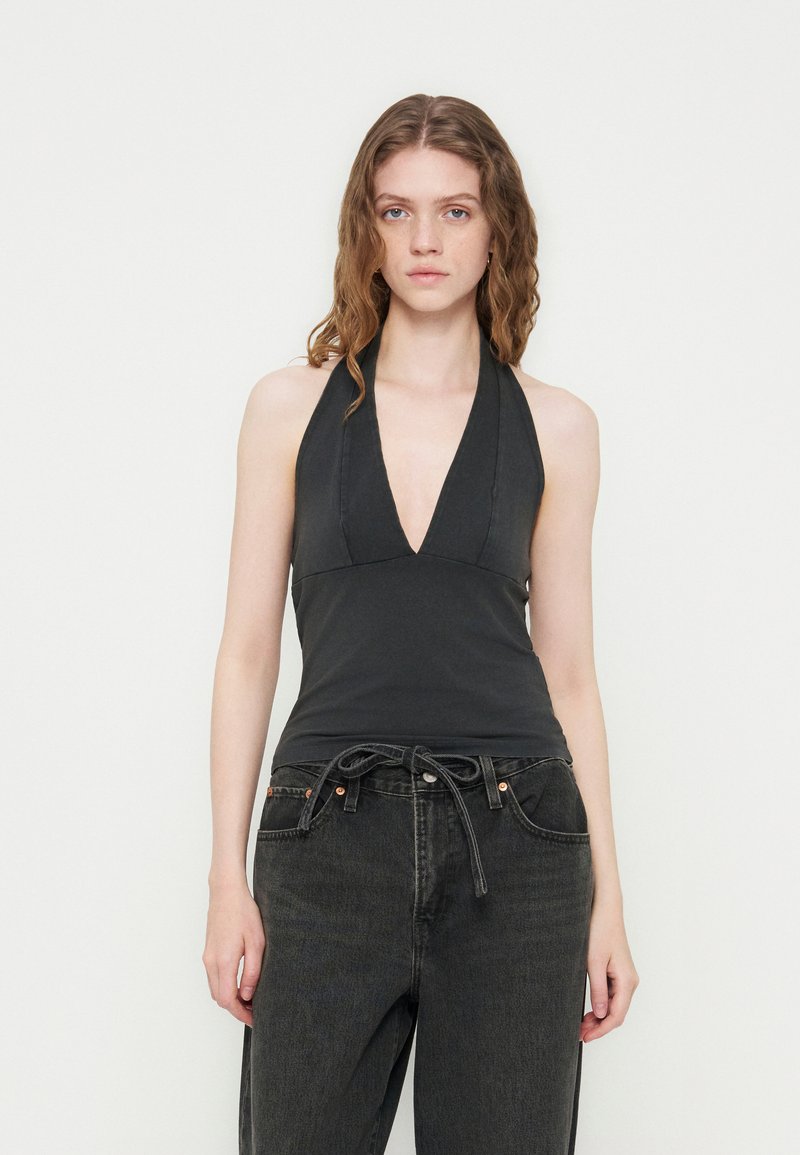 Jeune femme aux cheveux ondulés portant un haut noir sans manches à col halter et un jean large gris foncé avec une ceinture nouée.