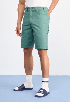 Mann trägt grüne knielange Shorts mit Seitentasche, weiße Socken und marineblaue Slides auf einem hellblauen Boden.
