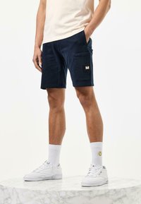 Námornícke modré cargo šortky s bočným vreckom, vyrobené z bavlny, s ležérnym strihom. V kombinácii s bielymi teniskami a krátkymi ponožkami s dizajnom usmievajúcej sa tváre.