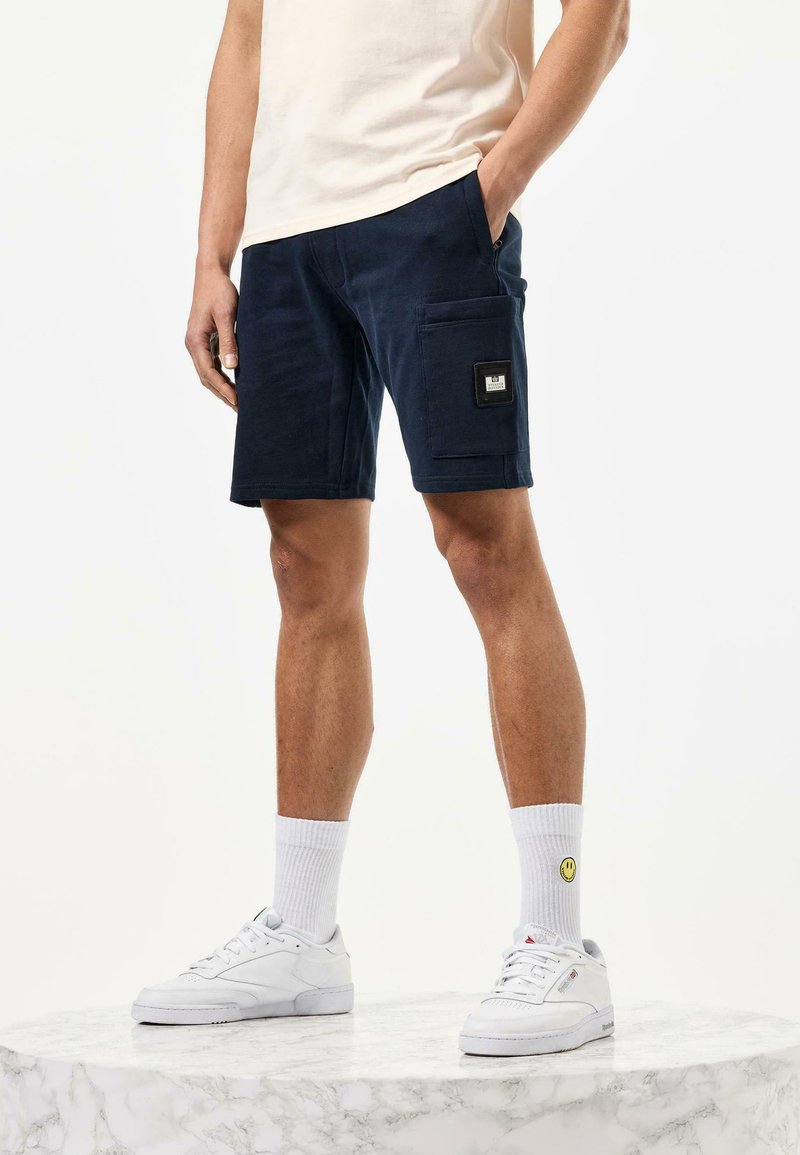 Námornícke modré cargo šortky s bočným vreckom, vyrobené z bavlny, s ležérnym strihom. V kombinácii s bielymi teniskami a krátkymi ponožkami s dizajnom usmievajúcej sa tváre.