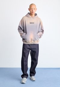 Light purple ombre hoodie with front pocket and printed logo, paired with dark denim pants and gray sneakers on a blue surface.
