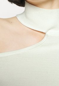 Ribbed knit top in off-white, featuring a high neck and cutout shoulder detail, highlighting smooth texture and form-fitting shape.