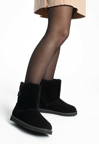 Botines de ante negro con forro de piel sintética, puntera redonda y suela de goma texturizada, combinados con medias negras transparentes y una falda beige.