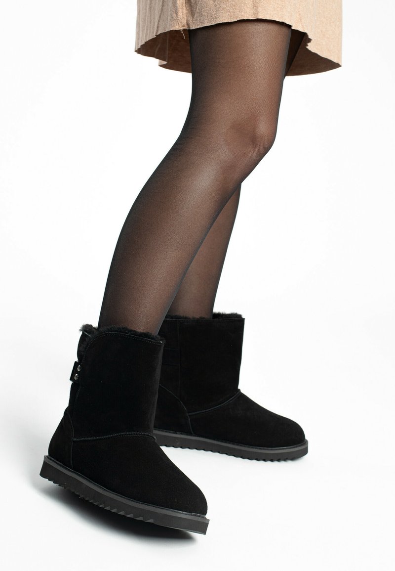 Botines de ante negro con forro de piel sintética, puntera redonda y suela de goma texturizada, combinados con medias negras transparentes y una falda beige.