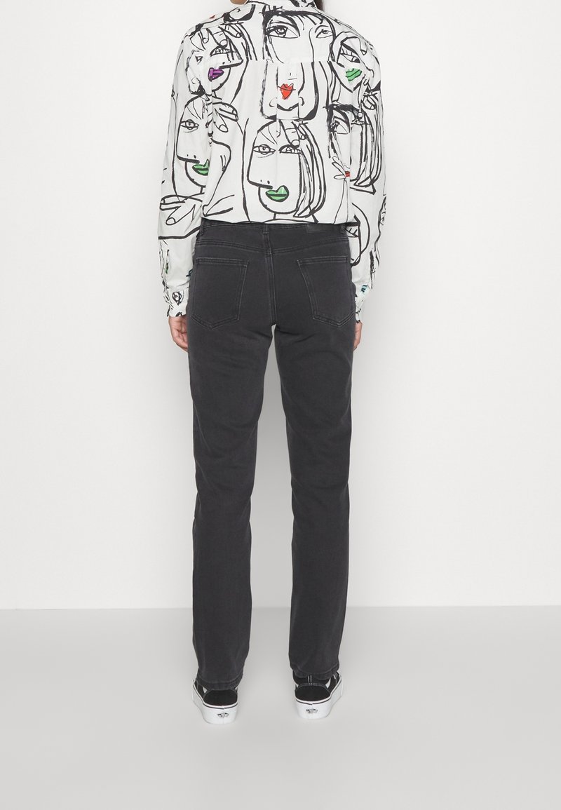 Personne portant un jean noir à jambe droite, des baskets noires et blanches, et une chemise à manches longues blanche avec un art de ligne abstrait représentant un visage en couleur.