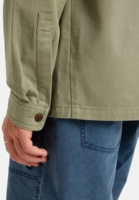 Olive green cotton shirt with a button closure at the cuff, paired with textured blue pants featuring side pockets and contrasting stitching.