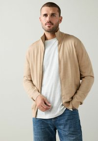 Kreminės spalvos zip-up kardiganas, pagamintas iš lygaus audinio, su ilgomis rankovėmis ir minkšta tekstūra, dėvimas ant baltos marškinės ir derinamas su mėlynomis džinsomis.