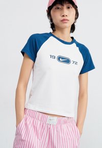 Valge puuvillane T-särk siniste raglanvarrukatega, millel on tumesinine Nike logo ja "1972" graafika. Koos roosade triibuliste lühikestega.