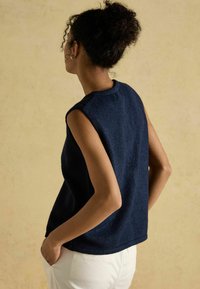 Jūras spēku tumši zils bezpiedurkņu tops no teksturēta auduma, ar apaļu kakla izgriezumu un brīvu piegriezumu, savienots ar gaišiem biksēm.