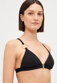 Svart trekant-bikini topp med teksturert stoff, gullsirkelaccent på stroppen og en solid kant under brystet for støtte.