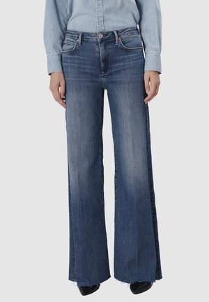 Femme portant un jean bleu taille haute à jambes larges avec des détails délavés, associé à une chemise en denim bleu clair rentrée à l'intérieur et des chaussures noires pointues.
