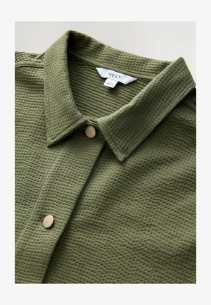 Olive green waffle-knit shirt with a pointed collar and a single round tan button at the front. Label reads “NEXT.” Soft texture.