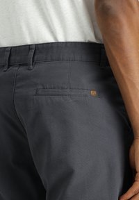 Chinos gris oscuro con una textura suave y un bolsillo trasero que presenta una pequeña etiqueta de marca marrón. La cintura tiene pasadores para cinturón.