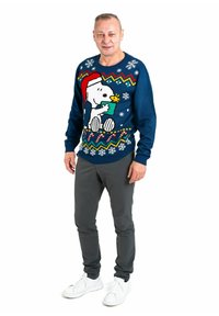 Navy blue sweater featuring Snoopy in a Santa hat, colorful pattern with zigzags and snowflakes, paired with gray pants and white sneakers.