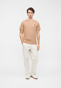 Ανοιχτό καφέ βαμβακερό t-shirt με κοντά μανίκια, με ένα μικρό κίτρινο λογότυπο πεταλούδας στο στήθος. Συνδυάζεται με φαρδιά μπεζ παντελόνια.