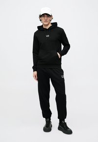 Czarna bluza z kapturem z przednią kieszenią i ściągaczem oraz czarne spodnie dresowe. Oba elementy wyglądają na wykonane z miękkiego materiału. Stylizację uzupełniają czarne trampki.