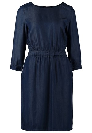 Femme portant une robe bleu marine longueur genou avec manches trois-quarts, collants noirs, bottines noires et un sac d'épaule bicolore.