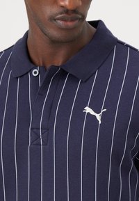 Mørkeblå polo shirt med hvide lodrette striber, knaplukket krave og broderet hvidt logo på brystet. Bomuldsstof med tekstureret finish.