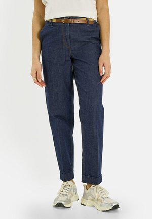 Persona con jeans azul oscuro con dobladillo, cinturón marrón, zapatillas blancas y camiseta blanca, de pie frente a un fondo blanco.