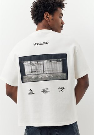 Mann trägt ein weißes T-Shirt mit einem schwarz-weißen Foto eines abgesperrten Ausgangs und Texten, darunter "Hollywood Boulevard" und "Full Time Vacation".