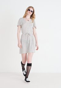 Abito a maniche corte a righe con cintura da annodare, realizzato in tessuto morbido. Indossato con collant neri e sneakers. Modella con occhiali da sole. Sfondo bianco.