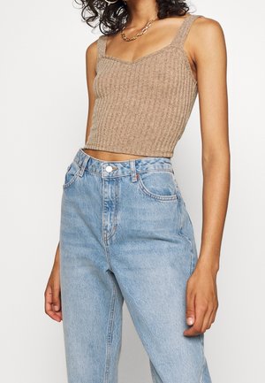 Vrouw die een mouwloze beige geribde cropped top en lichtblauwe high-waisted jeans draagt, gecombineerd met een gouden ketting en oorringen.
