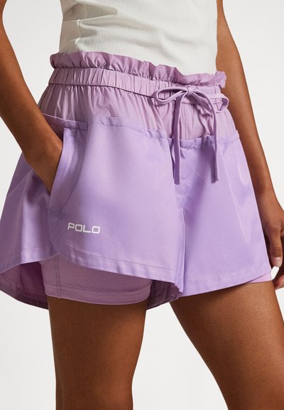 Γυναίκα που φοράει λιλά στρωματοποιημένα σορτς με ελαστική μέση και κορδόνι, με το ένα χέρι στην τσέπη, ορατό λογότυπο "POLO" στην αριστερή τσέπη.