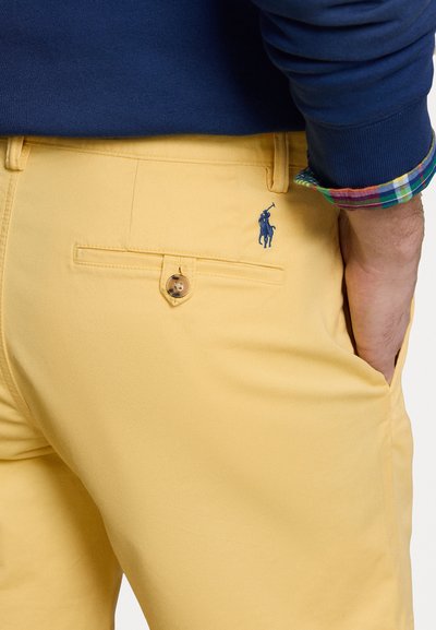 Calças de tecido amarelo com uma textura suave, apresentando um bolso traseiro com fecho de botão e um logótipo bordado azul.