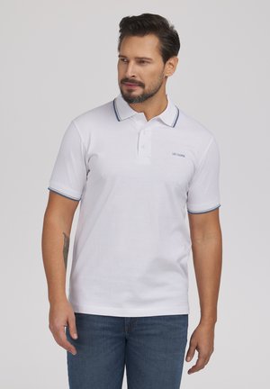 Mężczyzna w białej koszulce polo z niebieskim wykończeniem i logo "Lee Cooper", w połączeniu z niebieskimi dżinsami, stojący na jasnym tle.